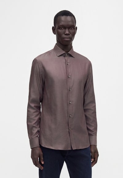 Άνδρας που φοράει μια μακρυμάνικη, κουμπωτή, καφέ γυαλιστερή πουκάμια με γιακά, στέκεται μπροστά σε ένα απλό λευκό φόντο.