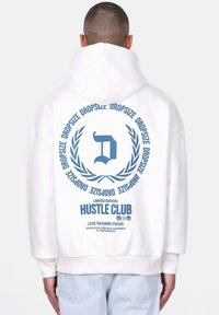 Bijela oversized dukserica s plavim grafičkim uzorkom na leđima, koja sadrži dizajn venca, slovo 'D' i tekst "LIMITED EDITION HUSTLE CLUB".