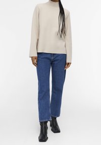 Beige coltrui met lange mouwen, gecombineerd met rechte blauwe jeans en zwarte enkellaarsjes met een vierkante neus.