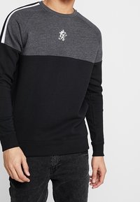 Maglione con metà superiore grigia e metà inferiore nera, con strisce bianche sulle maniche e un logo sul petto; realizzato in tessuto liscio.