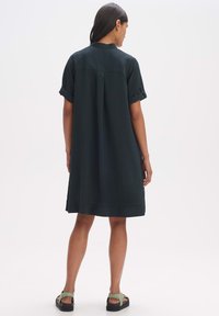 Dunkelgrünes, locker sitzendes Kleid mit kurzen aufgerollten Ärmeln, strukturiertem Stoff und knielangem Saum. Kombiniert mit schwarzen Sandalen und grünen Akzenten.