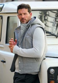 Mann in grauer Kapuzenweste und gestreiftem Pullover lehnt an einem weißen Geländewagen und schaut mit ernstem Blick in die Kamera.