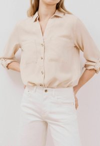 Camicia beige con bottoni, due tasche sul petto, maniche arrotolate e una consistenza morbida, abbinata a jeans bianchi dalla vestibilità classica.