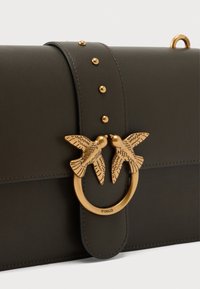 Σκούρο πράσινο δερμάτινο τσάντα χειρός με χρυσό κυκλικό κούμπωμα που απεικονίζει δύο πλεγμένα πουλιά και διακοσμητικά χρυσά καρφιά κατά μήκος του λουριού.