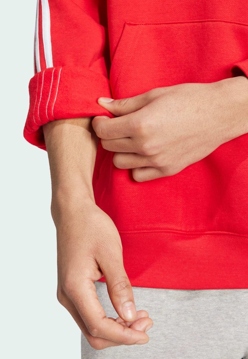 Sweat-shirt rouge avec des rayures blanches accents sur les manches retroussées. Mains ajustant la manche, mettant en valeur la texture côtelée et la poche kangourou.