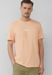Licht perzikkleurig T-shirt met korte mouwen, gemaakt van katoen, met een ronde halslijn en een klein wit geprinte logo op de borst. Zachte textuur.