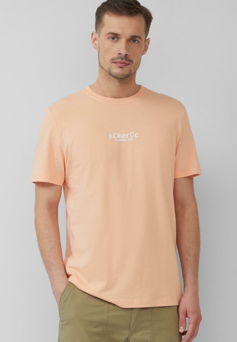 Licht perzikkleurig T-shirt met korte mouwen, gemaakt van katoen, met een ronde halslijn en een klein wit geprinte logo op de borst. Zachte textuur.