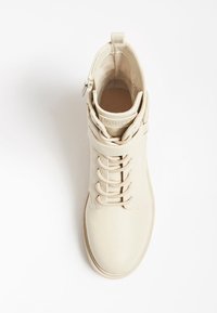 Bota beige de cuero al tobillo con cordones, textura suave, punta redondeada y correa decorativa en el tobillo. Diseñada para uso casual.