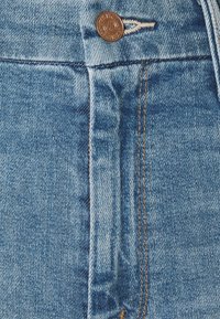 Primer plano de unos jeans de mezclilla azul con un botón visible, costura del cierre y detalles de costura naranja en la textura del tejido.
