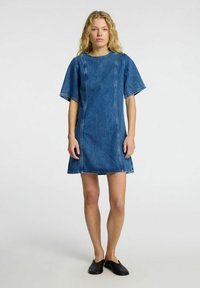 Blå denimshiftkjole med korte ermer og kontrastsøm. A-formet fasong og råkant. Svarte slip-on-sko brukt. Glatt tekstur.