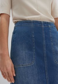 Jupe en denim à lavage moyen avec une taille haute. Elle possède deux poches avant et des coutures visibles. Matière en coton doux avec une légère texture.