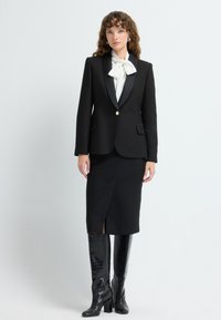 Blazer noir avec revers en satin, bouton doré, associé à une jupe crayon noire avec une fente avant. Bottes noires en cuir estampé crocodile arrivant au genou.