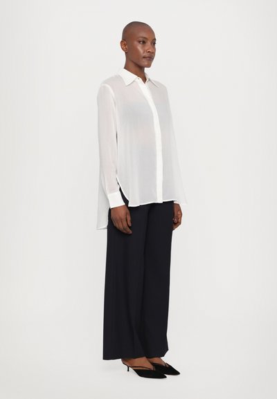 White, long-sleeve, button-up shirt with a collar, sheer fabric, side slits, worn with wide-leg, dark navy trousers and black heels.