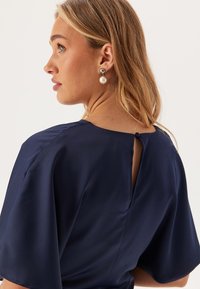Tmavě modrá blůza s krátkým rukávem, s kulatým výstřihem a malým zapínáním na knoflík na zadní straně. Hladká látka s jemným leskem.