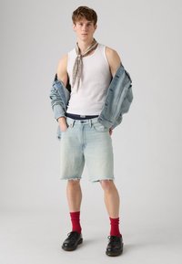 Veste en denim, débardeur blanc, shorts en denim bleu clair avec un ourlet effiloché, chaussettes rouges et chaussures noires. Écharpe à motifs autour du cou.