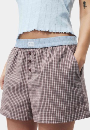 Person trägt lockere karierte Shorts mit blauem elastischem Bund und drei Knöpfen, kombiniert mit einem hellblauen bauchfreien Oberteil, das ein Tattoo im mittleren Bauchbereich zeigt.