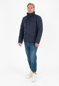 Giacca invernale blu navy con colletto alto, numerose tasche frontali e una toppa con logo. Indossata sopra jeans azzurri e sneakers bianche.