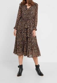 Femme portant une robe midi à manches longues imprimée léopard avec une ceinture à la taille, associée à des bottines noires sur un fond uni.