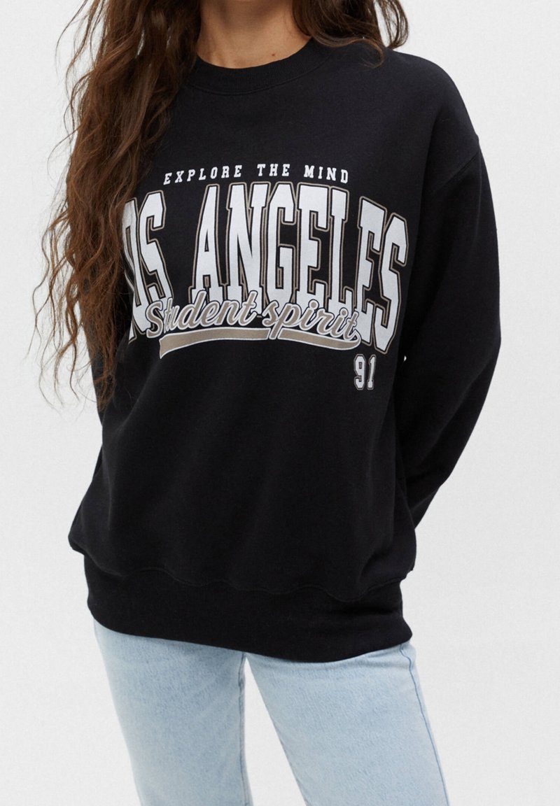 Femme portant un sweat-shirt noir avec un texte blanc « Explore the Mind Los Angeles Student Spirit 91 » et un jean bleu clair.