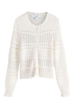 Kvinde med langt mørkt hår står med hænderne i lommerne iført en brun jakke, hvid tekstureret top, brede beige bukser og leopardmønstrede flade sko.