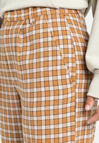 Gecontroleerde broek in oranje en witte stof, met een hoge taille, plooien en zijzakken. De textuur lijkt zacht en gestructureerd.