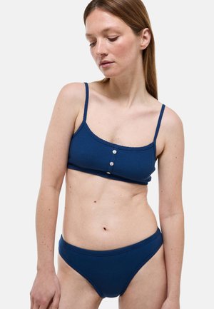 Jeune femme aux cheveux raides châtain rouge portant un bralette côtelé bleu marine avec des boutons et une culotte taille haute assortie.