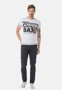 Weißes T-Shirt mit kurzen Ärmeln und schwarzem Grafikdruck, mit "CIPO & BAXX". Kombiniert mit dunkelblauen Hosen und weißen Sneakern.