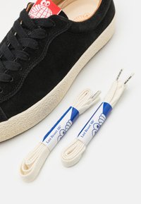 Schwarze Wildledersneaker mit strukturiertem Obermaterial, weißer Gummisohle und schwarzen Schnürsenkeln. Enthält zwei cremefarbene Ersatzschnürsenkel in blauer Verpackung.
