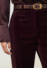 Bordo baršunaste hlače s glatkim bočnim džepovima, uparene s smešanim pojasom s uzorkom koji ima zlatnu kopču, djelomično vidljiva rukava.