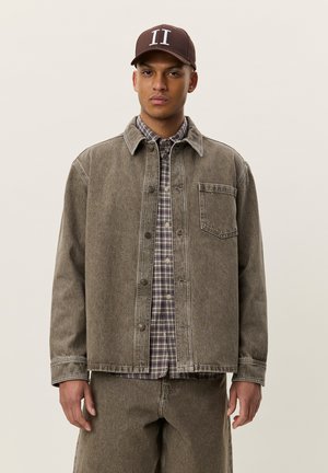 Ung mand iført brun baseballkasket, brun falmet denimjakke over ternet skjorte og matchende denimbukser, stående mod en ensfarvet baggrund.