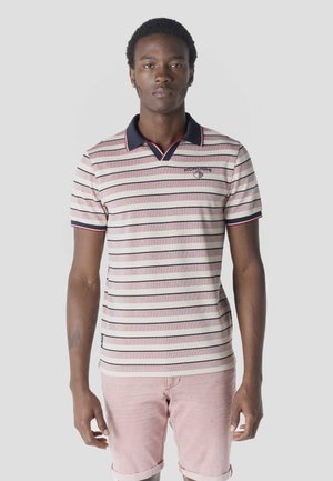 Mladý muž stojaci, oblečený v pruhovanom polokošeli s námorníckym golierom a ružovými šortkami, tvárou smerujúci dopredu na jednoduchom pozadí.