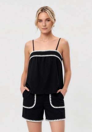 Lyshåret kvinde iført sort ærmeløs top og shorts med hvid trekantet kant, stående med hænderne i lommerne mod grå baggrund.