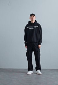 Schwarzer Hoodie mit dem Text "DSQUARED2 HERITAGE Established 1995", kombiniert mit schwarzen Cargo-Hosen und weißen Sneakers. Einfaches, urbanes Design.