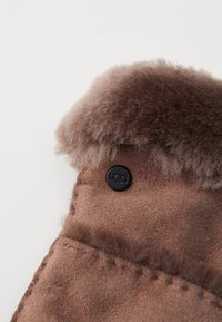 Brune skinnhanske med myk tekstur, med en fluffy mansjett og en svart UGG-logo knapp. Brodert detaljer langs kanten.