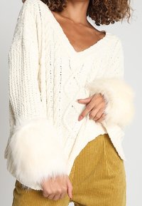 Pull en tricot ivoire avec col en V, motif torsadé et poignets en fausse fourrure douce. Associé à un pantalon en velours côtelé couleur moutarde.