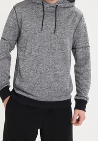 Mężczyzna w szarym, melanżowym hoodie z czarnymi sznurkami i czarnymi mankietami, w połączeniu z czarnymi spodniami, stojący z luźno opuszczonymi rękami.