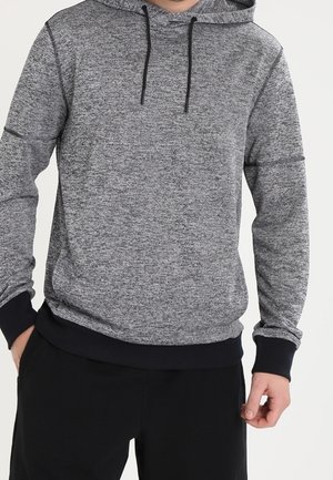 Man i en grå melerat hoodie med svarta snören och svarta mudd, tillsammans med svarta byxor, stående med avslappnade händer.