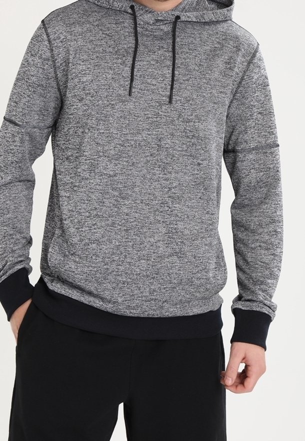 Mężczyzna w szarym, melanżowym hoodie z czarnymi sznurkami i czarnymi mankietami, w połączeniu z czarnymi spodniami, stojący z luźno opuszczonymi rękami.