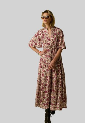 Femme en robe maxi à fleurs avec manches courtes, lunettes de soleil surdimensionnées et bottines noires, debout avec une main sur la hanche.