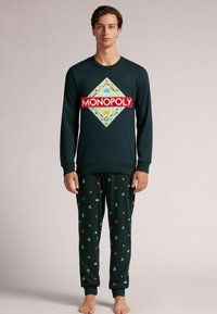Sudadera verde de manga larga con el logo rojo de "MONOPOLY" y diseño de diamante, combinada con pantalones de pijama verdes que presentan acentos de patrón coloridos.