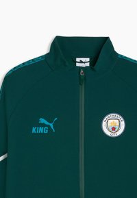 Veste de sport zippée vert foncé avec logo Puma et texte "KING" sur la poitrine gauche, blason de Manchester City sur la poitrine droite, bandes d'épaule bleues à motif.