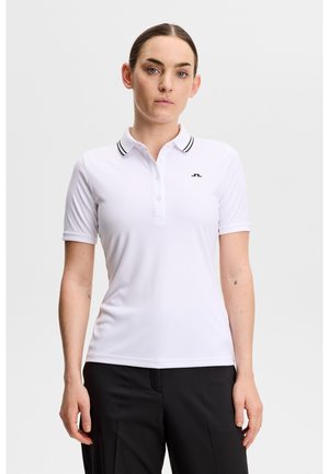 Vrouw draagt een wit poloshirt met korte mouwen met zwarte bies aan de kraag en een zwarte broek, staand tegen een effen achtergrond.