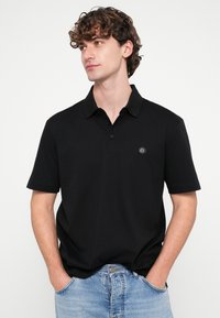 Melna polo krekls ar īsām piedurknēm, apkakli un priekšpuses pogu aizdari. Kreisajā krūšu daļā ir neliels logotips. Gluda auduma tekstūra.