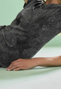 Grå T-shirt med en tekstureret finish, der har sorte glitrende stjerne-mønstre. Rund halsudskæring og korte ærmer, lavet af blødt stof.