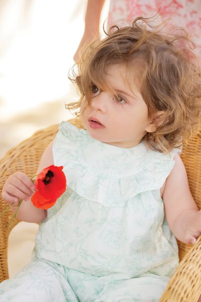 Light green floral romper with ruffled collar, made of soft fabric. Child holds a bright red poppy flower, seated in a wicker chair.