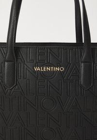 Fekete bőr bevásárlótáska, a "VALENTINO" logo dombornyomott, ismétlődő mintázatával és arany színű fémrészekkel a márkanevében.