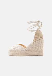 Espadrille compensée beige avec plateforme tissée, bout en dentelle florale, arrière fermé et liens à nouer à la cheville ornés de pompons sur fond blanc.