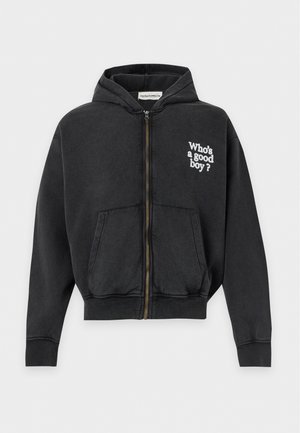 Homme portant un sweat à capuche noir avec deux chiens dessinés et le texte "Who's a good boy?" dans le dos, associé à un pantalon noir et des chaussures.