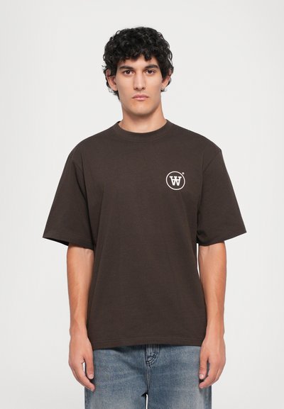 Uomo con capelli ricci che indossa una maglietta oversize marrone scuro con un piccolo logo circolare bianco sul petto, abbinata a jeans blu.