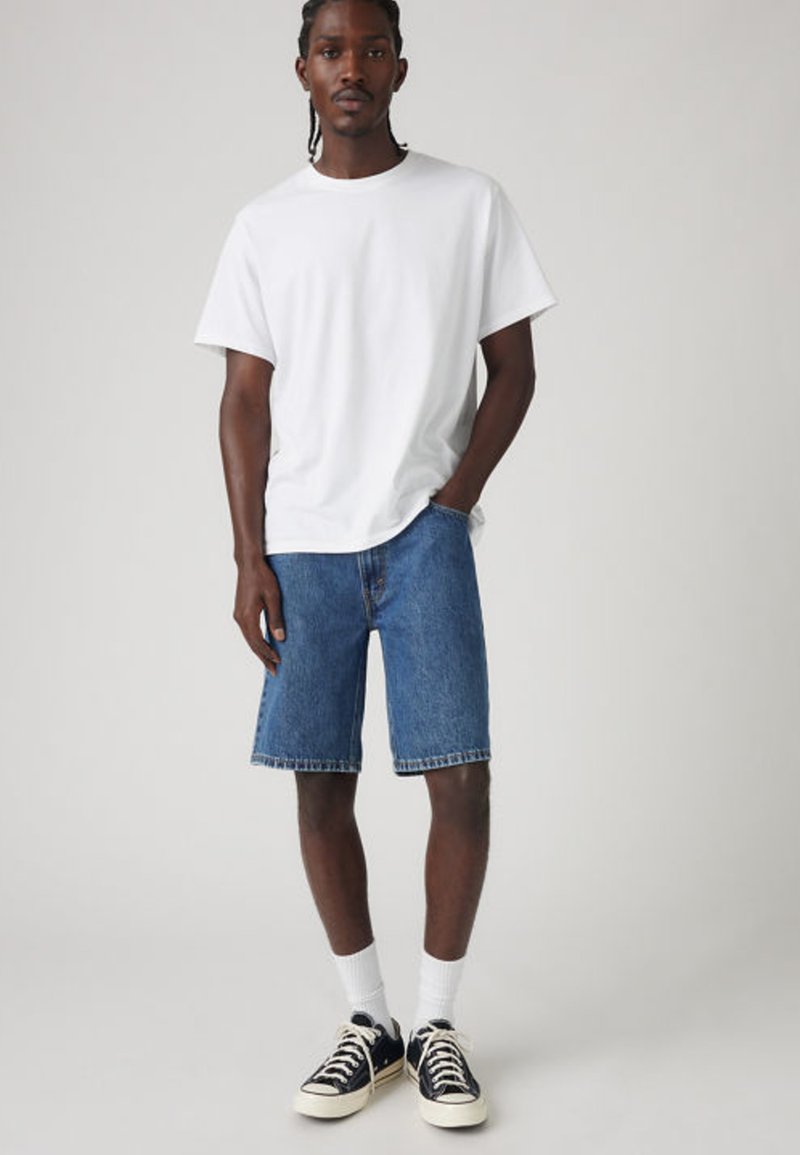 Homme aux cheveux tressés portant un t-shirt blanc, un short en denim bleu, des chaussettes blanches et des baskets noires, debout devant un fond uni.